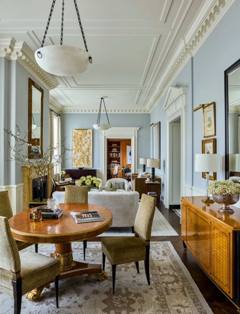 Elegant living-dining room in a luxury Fifth Avenue residence, featuring ornate moldings, high ceilings, light blue walls, and large mirrors. A round wooden table with books, green chairs, beige sofas, floral arrangements, sideboard, and artwork create a refined, classic atmosphere. Light floods from windows.