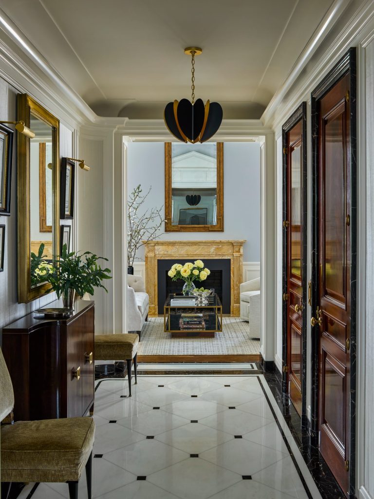 Elegant Fifth Avenue residence featuring a hallway with a black and white marble floor, gold-framed mirrors, and wood doors. The hallway leads to a living room with a gold fireplace, large mirror, white sofas, coffee table topped with yellow flowers, and a branch in a vase. A black modern chandelier hangs above.