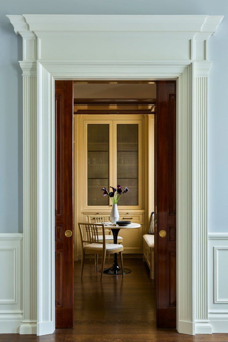 A small dining nook in this Fifth Avenue residence features a round table, two wooden chairs, and a built-in bench, all viewed through open wooden pocket doors. A white vase with dark purple tulips sits on the table. The walls are light blue with white trim and paneling.