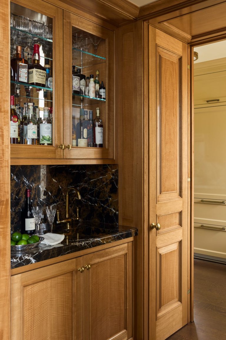 A small home bar in this elegant Fifth Avenue residence features wood cabinets, glass shelves holding liquor bottles and glassware, a dark marble countertop and backsplash, and a brass faucet. Limes and a knife rest on a plate, while a wooden door stands open to reveal a cream refrigerator in the next room.