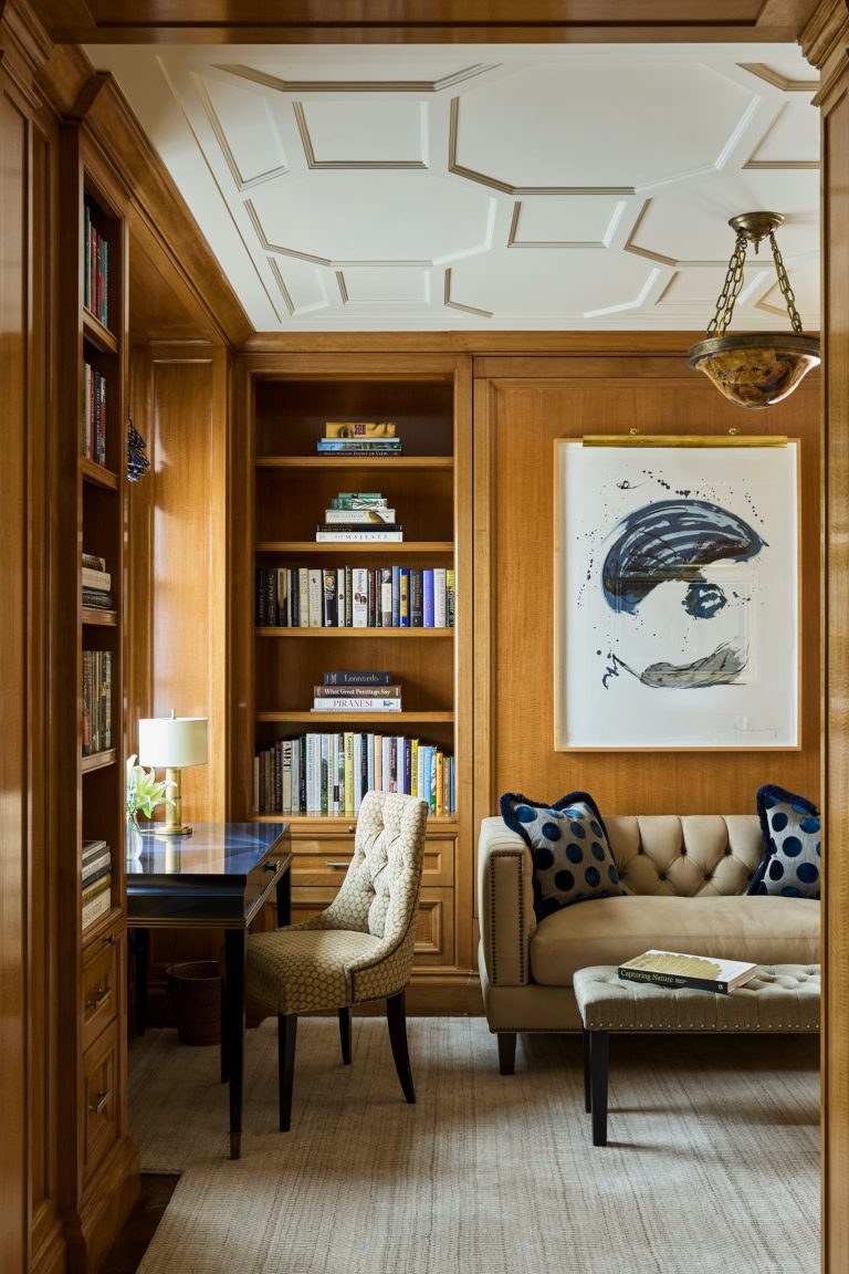 A cozy wood-paneled study in a Fifth Avenue residence, with bookshelves, abstract art, a tufted beige chair at a black desk with a lamp, and a beige couch adorned with blue polka dot pillows. A coffee table, rug, and ceiling moldings add elegance while warm, natural lighting fills the room.