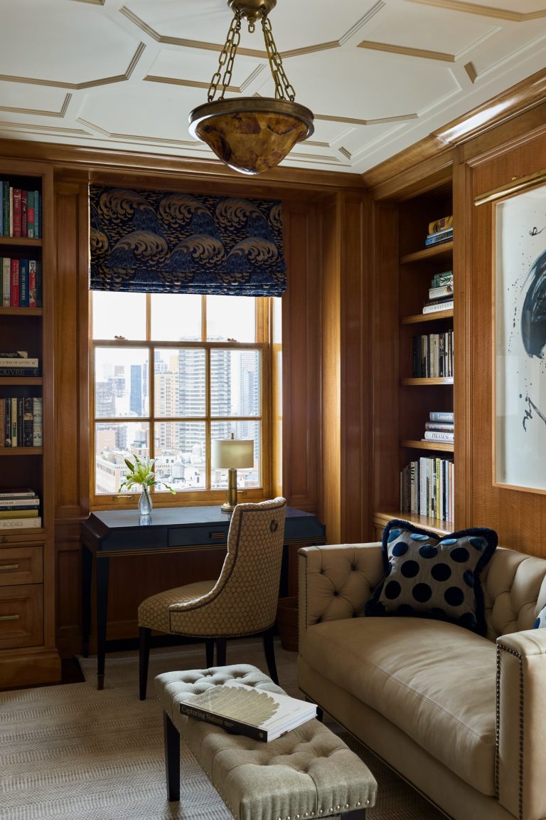 Elegant wood-paneled study in a Fifth Avenue residence featuring a tufted beige sofa, polka-dot pillow, matching ottoman, and desk with chair by a window offering a stunning city view. Built-in bookshelves, abstract art, a lamp, and a floral arrangement complete the cozy, sophisticated space.