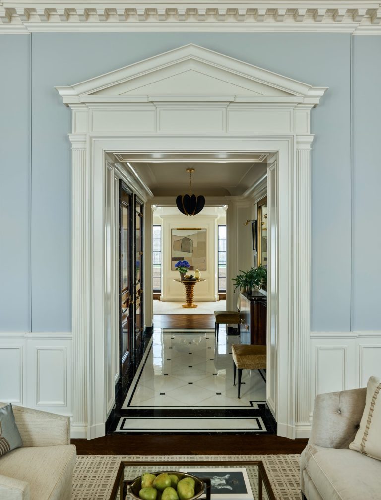 A hallway in this elegant Fifth Avenue residence, with white trim, marble floor, and classical molding, leads to a room where a round table holds a vase of yellow flowers. Art and windows are visible, furniture and green decor accent the space. Two beige chairs frame the foreground.