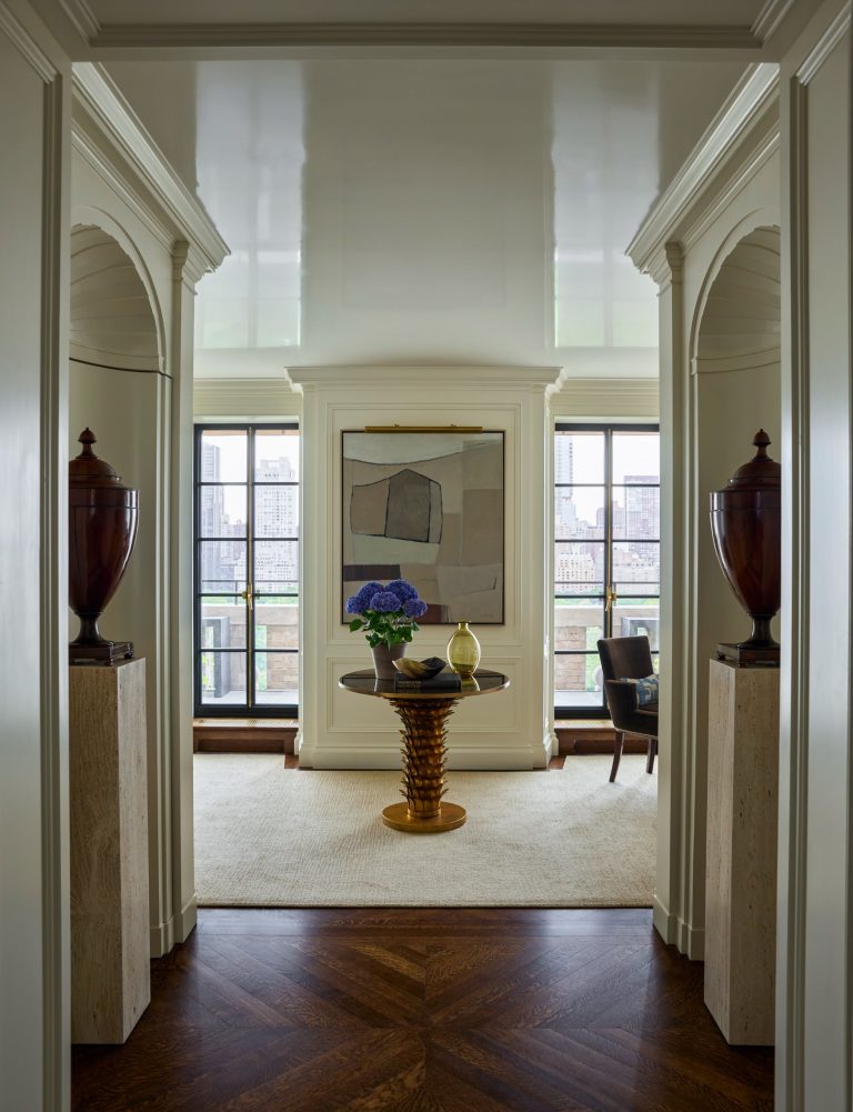 A symmetrical hallway leads to a room with two large urns on pedestals, a round table with blue flowers, modern art on the wall, large windows letting in daylight, and a lounge chair to the right. This elegant Fifth Avenue residence is defined by neutral tones, geometric flooring, and an inviting sense of sophistication.