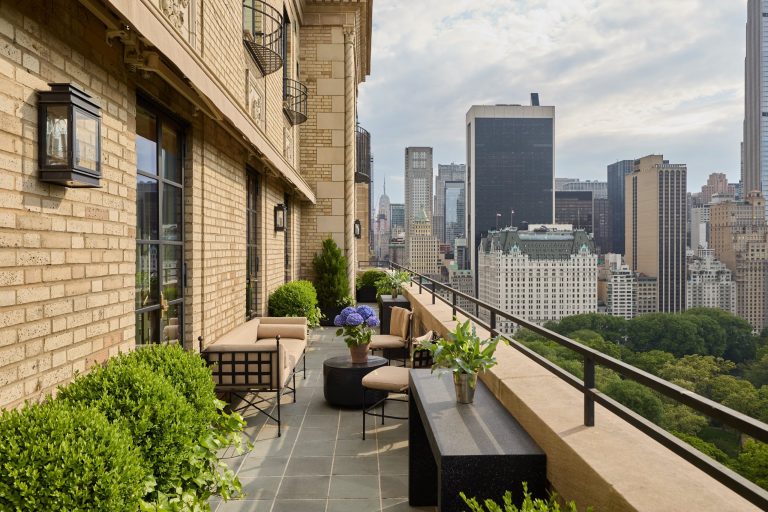 A stylish Residence on Fifth Avenue balcony with beige sofas, armchairs, potted plants, and small tables overlooks a cityscape of tall buildings and Central Park. The sky is partly cloudy, and greenery from the park contrasts with the urban skyline in the background.