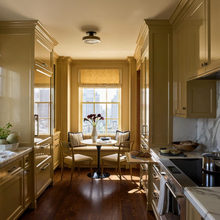 Narrow, elegant kitchen with glossy beige cabinets, marble backsplash, and wood floors. At the sunlit window of this sophisticated Fifth Avenue residence, a round table with two striped chairs and a vase of dark flowers creates an inviting nook. City buildings are visible outside as sunlight streams in, creating a warm, cozy atmosphere.