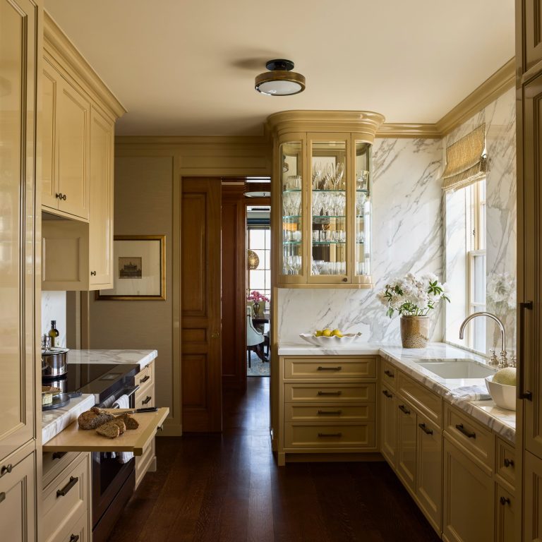 Elegant Fifth Avenue residence kitchen with cream cabinets, marble countertops, and backsplash. Glass-front upper cabinet holds glassware. Sink below window with flowers. Dark wood floor, tray of bread on oven pull-out shelf, lemons on counter. Artwork and doorway visible in background.