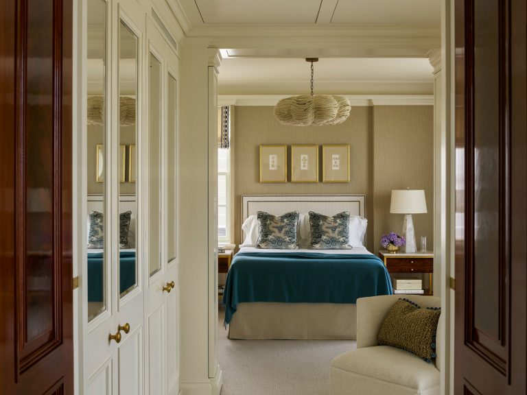Elegant bedroom in the Residence on Fifth Avenue with a bed featuring blue and white pillows, teal blanket, wood nightstands with lamps, framed art above the headboard, beige walls, mirrored closet doors, and an armchair with a textured pillow in the foreground.