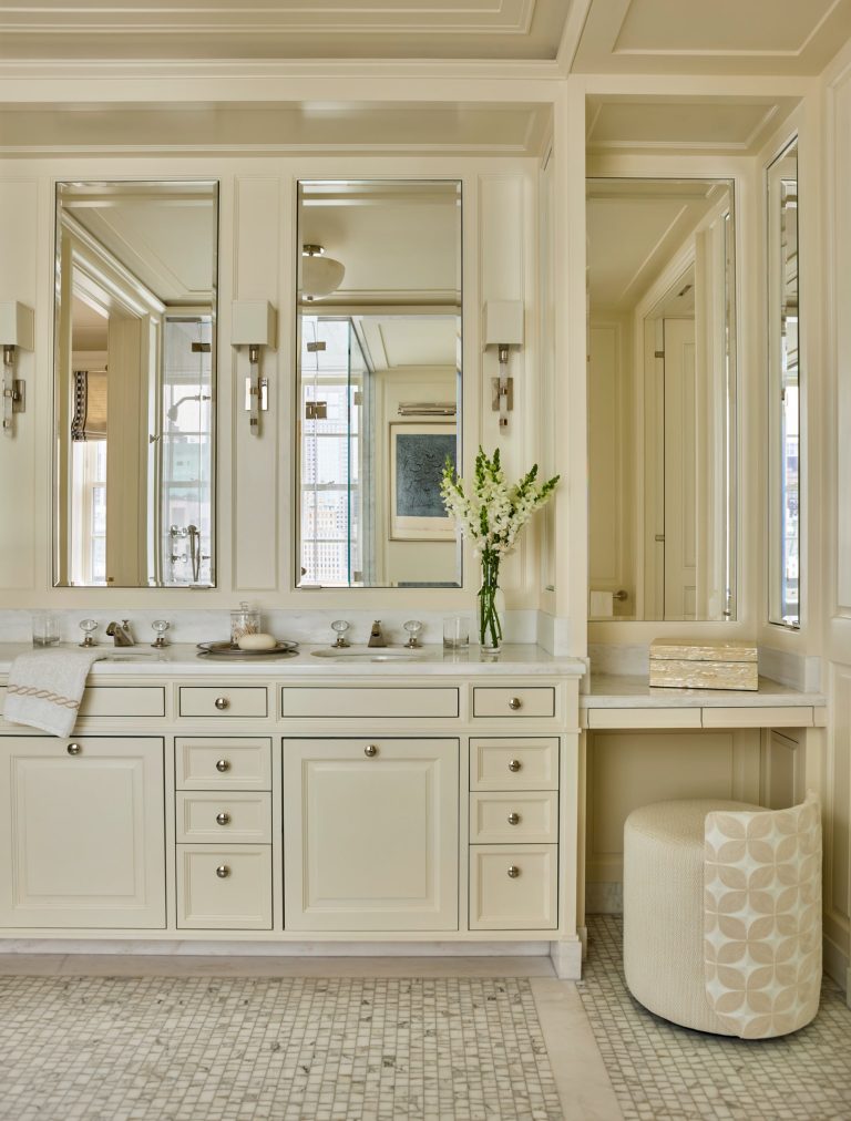 Elegant bathroom in a Fifth Avenue residence with cream cabinetry, double sinks, and three tall mirrors. Silver fixtures, wall sconces, and a white vase with flowers sit on the marble countertop. A vanity area includes a cushioned stool and a decorative box. The floor has small, square tiles.