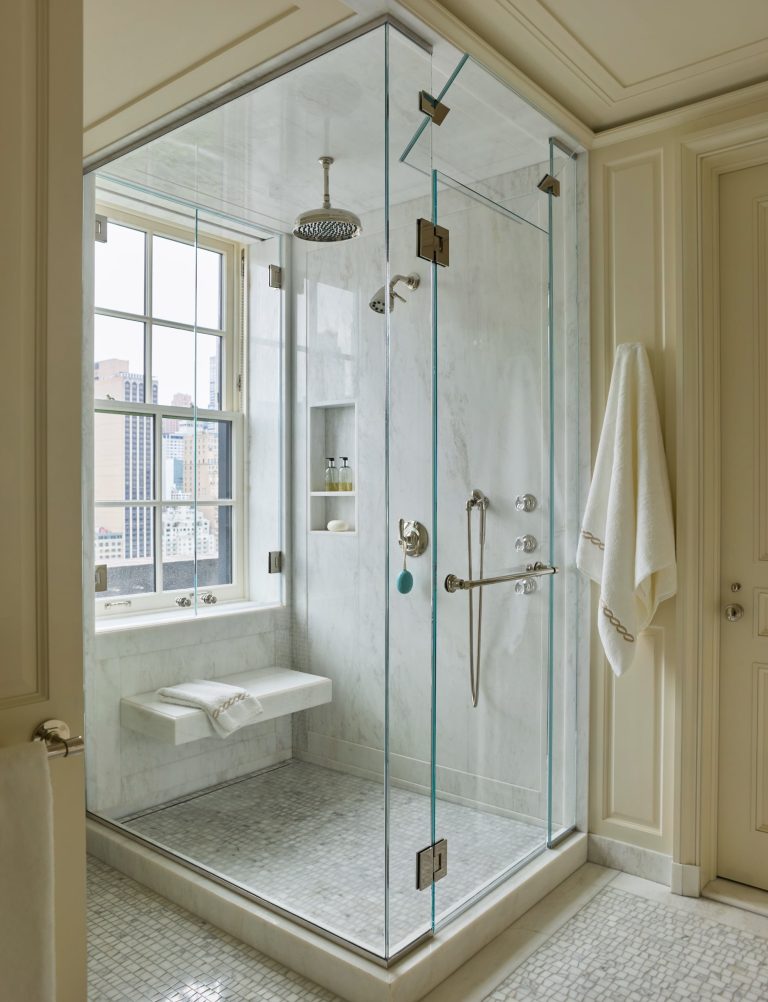 A modern glass shower with marble walls, a rain showerhead, a wall-mounted bench, built-in shelves with toiletries, silver fixtures, and a white towel hanging outside. Sunlight streams through a large window, revealing a stunning Fifth Avenue cityscape view—an elegant retreat in an exclusive residence.