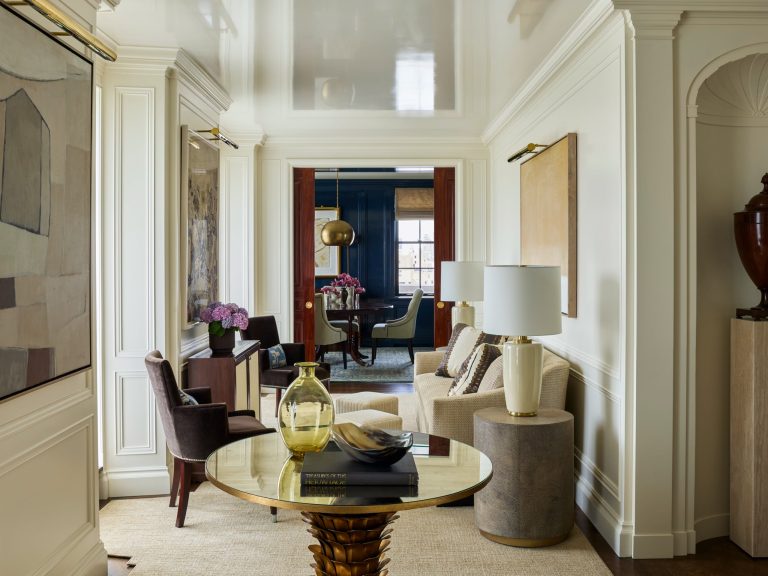 Elegant living room with cream walls, modern art, plush chairs, a round gold table with books and a glass vase, a sofa, table lamps, and purple flowers. This Fifth Avenue residence exudes luxury throughout; in the background, a dining area features striking blue walls, a gold light fixture, and a window.