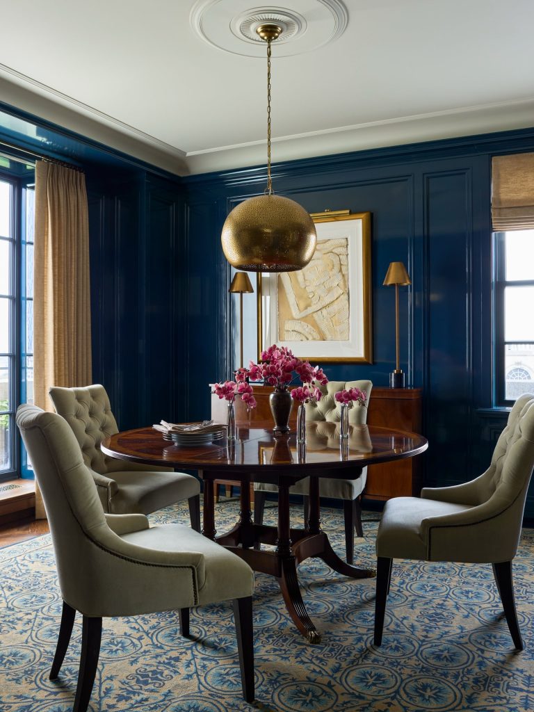 Elegant dining room in a Fifth Avenue residence with dark blue paneled walls, a round wooden table with four tufted chairs, a gold pendant light, and pink flowers in vases. Large windows with tan curtains illuminate the space, and a blue patterned rug covers the floor.