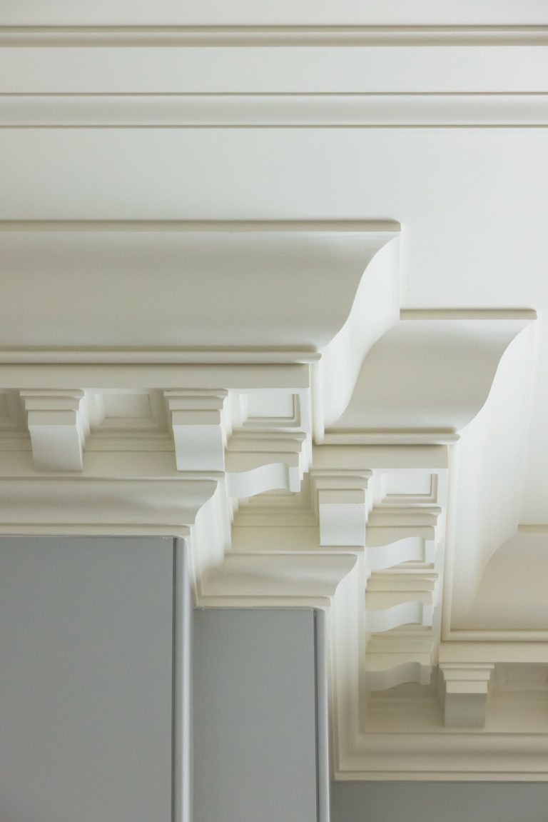 Detailed white crown molding decorates the ceiling of this luxury apartment, featuring intricate, layered designs and deep shadow lines. The molding contrasts with the smooth gray and white walls below, highlighting the architectural detail and craftsmanship of this Fifth Avenue residence.