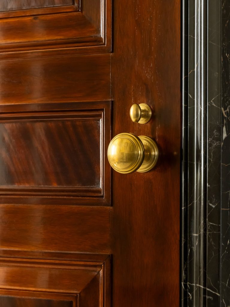 A close-up of a polished wooden door with a shiny brass doorknob and lock. The door is partially open, revealing a black marble doorframe—an inviting entrance to an elegant Fifth Avenue residence. The rich wood grain and reflective brass fixtures add an exquisite touch to the overall appearance.