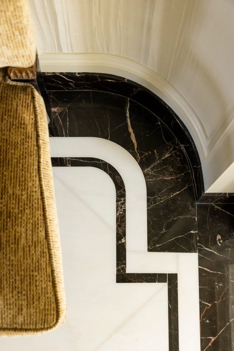 Curved corner of a Fifth Avenue residence with a sleek black and white marble floor featuring geometric patterns. Part of a textured beige upholstered chair is visible along the left edge. Walls are white with decorative trim. The scene is lit with soft natural light.