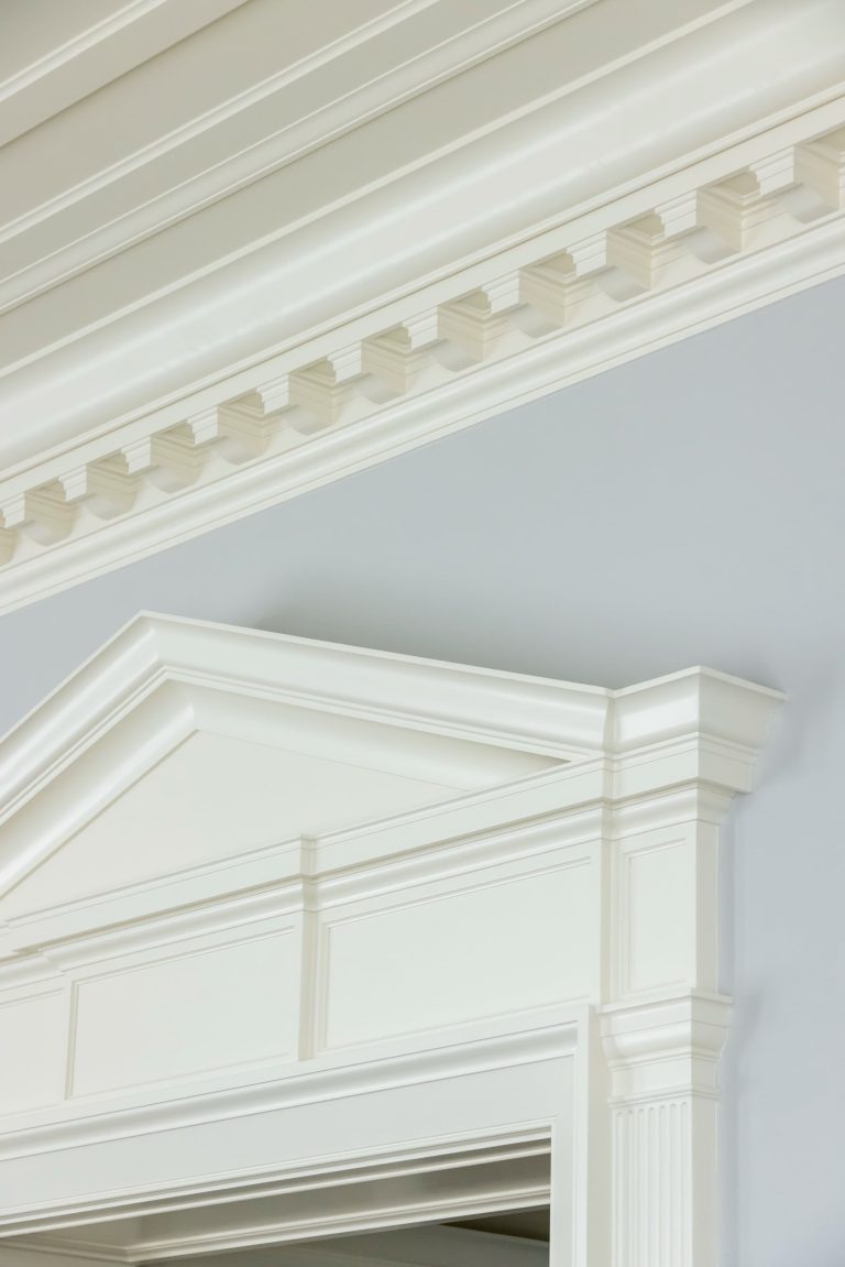 White crown molding and decorative trim frame a doorway and run along a light gray wall and ceiling, adding timeless elegance reminiscent of a Fifth Avenue residence. The classic architectural details include dentil molding, a peaked pediment above the doorway, and fluted vertical side pieces.